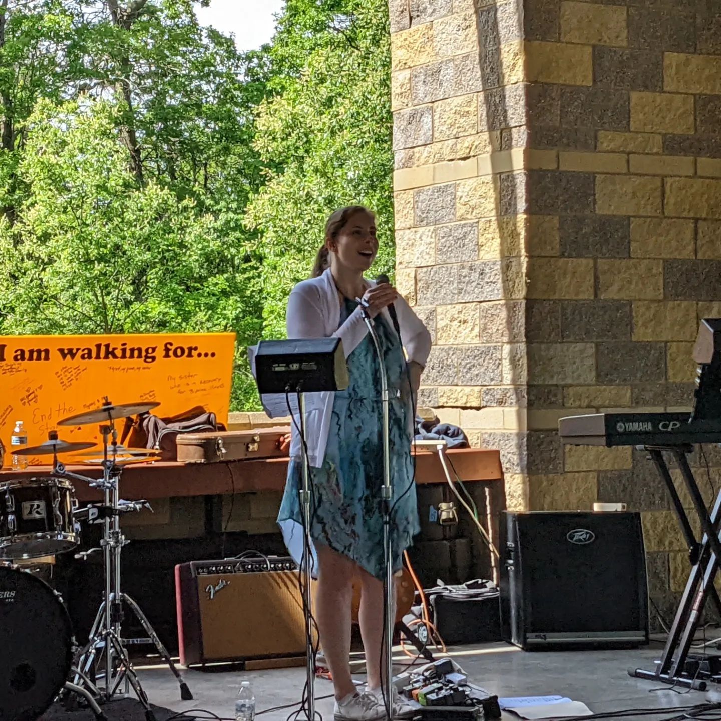 Sierra in a blue dress and a white cardigan with a microphone in her hand speaking in an outdoor area.